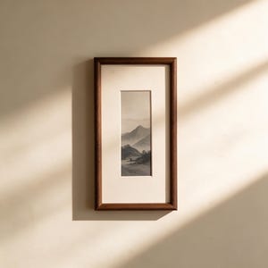 Puede incluir: Una pieza de arte mural rectangular vertical que presenta una ilustración de paisaje monocromático de montañas y árboles. La obra está encerrada en un marco de madera marrón oscuro con un paspartú blanco roto que rodea la impresión central.