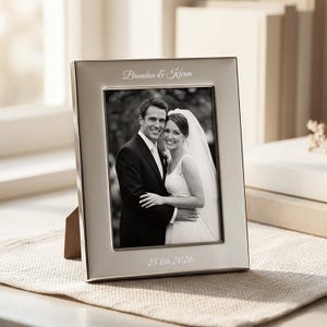 May include: A rectangular silver-toned metal picture frame featuring an engraved inscription at the top reading Brandon & Karen and a date reading 25.06.2026 at the bottom. The frame holds a black and white photograph of a couple in wedding attire.