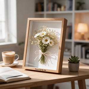 May include: A rectangular light wood shadow box frame containing a pressed bouquet of white daisies and small yellow filler flowers tied with a thin twine bow. The bottom frame edge features the engraved text Forever By My Side.