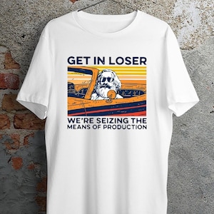 May include: A white short-sleeved t-shirt featuring a graphic print of a bearded figure in sunglasses driving an orange convertible car. The text reads GET IN LOSER and WE'RE SEIZING THE MEANS OF PRODUCTION, set against a sunset background.