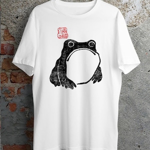 Könnte beinhalten: Ein weißes kurzärmeliges T-Shirt mit einer schwarzen Illustration im Tusche-Stil, die einen Frosch in der Mitte der Brust zeigt. Über dem Frosch befindet sich ein kleiner roter quadratischer Stempel mit asiatischen Schriftzeichen.