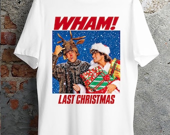 May include: A white short-sleeved t-shirt featuring a graphic print of the band Wham! with the text "WHAM!" and "LAST CHRISTMAS" in red. The image shows two individuals, one wearing a reindeer headpiece and the other a Santa hat with gifts.