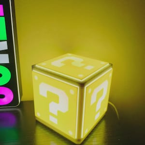 May include: A yellow cube-shaped lamp featuring a white question mark symbol on each visible side. The lamp emits a soft, warm glow, highlighting the square design and the distinct question mark graphics printed on the top and side panels.