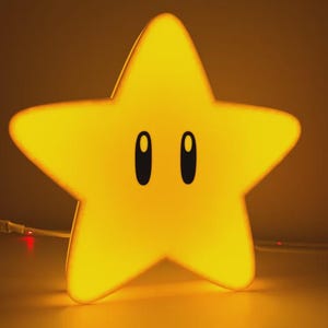 May include: A bright yellow star-shaped lamp with a smooth surface and two large, vertical black oval eyes centred on the front. The lamp emits a warm, golden glow and is connected to a thin white power cord with a small control switch attached.