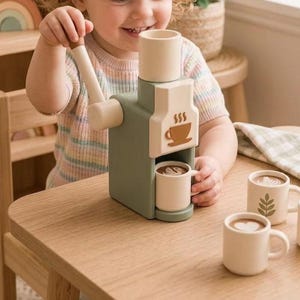 Puede incluir: Una máquina de café expreso de juguete en madera color verde salvia y crema, con manivela lateral, tolva superior y un panel frontal con un icono de taza de café marrón. El set incluye tres tazas pequeñas color crema con diseños de arte latte.