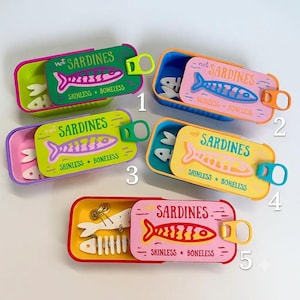 May include: Five rectangular metal tins with sliding lids, each featuring a fish graphic and the text "not SARDINES" or "SARDINES" with "SKINLESS + BONELESS". The tins are green, pink, lime, yellow, and red, containing white fish bone ornaments.