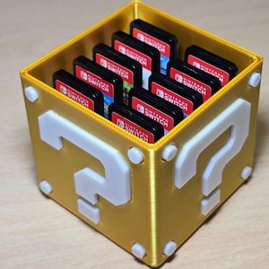 May include: A gold-coloured, cube-shaped storage container featuring white question mark symbols on its sides. The container is filled with multiple Nintendo Switch game cartridges, each displaying the Nintendo Switch logo on its label.