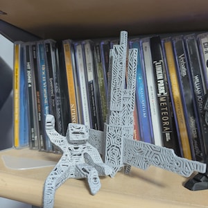 May include: A light gray 3D printed figure with a complex geometric lattice pattern. The figure features a blocky head, articulated limbs, and a tall, rectangular structure extending from its back, all covered in a repeating hexagonal design.
