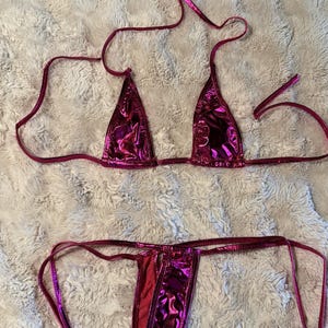 May include: A two-piece bikini set in a metallic hot pink finish. The top features triangular cups with thin adjustable tie straps. The bottom is a thong style with thin side ties, crafted from a shiny, reflective material with a smooth texture.