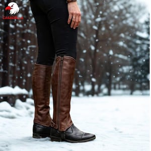 May include: Brown leather chaps worn over black pants and black boots. The chaps feature a full-length vertical zipper along the outer side, extending from the top edge down to the ankle area, providing a secure fit over the lower leg.