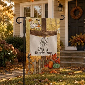 Puede incluir: Una bandera de jardín rectangular con una cruz de madera, dos gansos con sombreros y bufandas, calabazas y una valla de madera. El texto dice Fall for Jesus He never leaves. El fondo muestra un bosque otoñal con hojas naranjas.