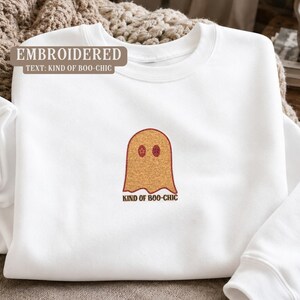 Peut inclure: Un sweat-shirt blanc à col rond avec une broderie centrale représentant un fantôme orange doré aux yeux circulaires rouge foncé. Sous le fantôme, le texte "KIND OF BOO-CHIC" est brodé en marron foncé sur la poitrine du vêtement.