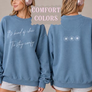 May include: A blue crewneck sweatshirt featuring the white text It's kind of chic To stay comfy on the front and three small white daisy flower outlines on the back. The garment is labelled as Comfort Colors in the blue jean colour shade.