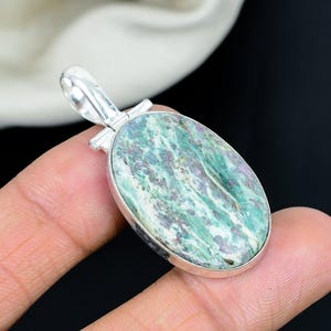 Puede incluir: Un colgante de forma ovalada con una piedra pulida multicolor que presenta patrones en espiral de color verde salvia, blanco y magenta intenso. La piedra está engastada en un marco de metal plateado liso y pulido con una anilla a juego.