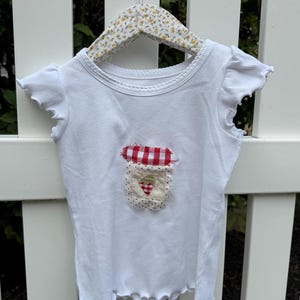May include: A white ribbed t-shirt with short ruffled sleeves and a round neckline. The center features a fabric appliqué of a jar with a red and white checkered lid and a small strawberry motif on a cream-colored background with frayed edges.