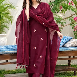 May include: A deep burgundy V-neck kurta set featuring small white and red floral embroidery scattered across the fabric. The outfit includes a matching pair of straight-leg trousers and a sheer, lightweight dupatta with tassel accents.