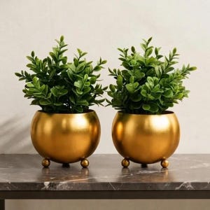 May include: Two identical spherical gold-coloured metal planters, each supported by three small ball-shaped feet. Each planter contains a lush, vibrant green artificial plant with small, rounded leaves that extend upwards and outwards from the centre.
