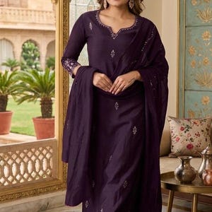 Könnte beinhalten: Ein dunkelviolettes Salwar-Kameez-Set, bestehend aus einer langärmeligen Tunika mit gekerbtem Ausschnitt, einer passenden Hose mit geradem Bein und einem Dupatta. Das Kleidungsstück ist mit zarter Silberstickerei am Ausschnitt und an den Ärmelbündchen verziert.