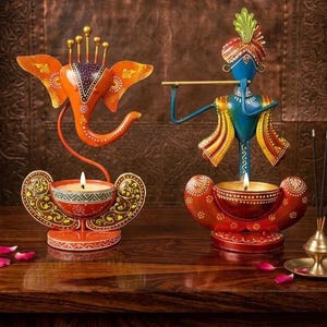 May include: Two metal candle holders featuring stylised figures. On the left, an orange elephant-headed figure with a purple forehead. On the right, a blue figure playing a gold flute. Both figures stand above circular bowls holding lit candles.