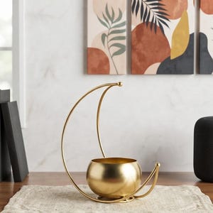 May include: A gold-colored metal decorative bowl featuring a crescent moon-shaped frame. The bowl is spherical with a smooth, reflective finish, and the thin, curved metal structure extends upward and around the base to create a moon silhouette.
