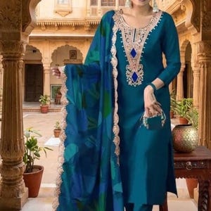 May include: A teal long-sleeved tunic and matching trousers set featuring an intricate beige embroidered neckline with blue diamond-shaped accents. A coordinating teal shawl with a blue and green leaf pattern and a scalloped beige border is draped.