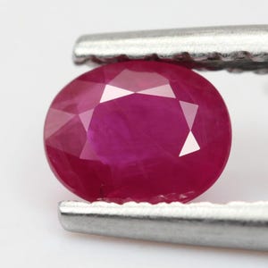 May include: An oval-cut, vibrant red ruby gemstone held by silver-toned metal tweezers. The gemstone features a faceted surface that reflects light, showcasing its deep crimson colour and internal clarity characteristics within the stone structure.