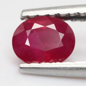 Op de afbeelding: Een levendige, ovaal geslepen robijn met een dieprode kleur en een gefacetteerd oppervlak. De edelsteen wordt vastgehouden door een zilverkleurige metalen pincet, wat de gepolijste helderheid en symmetrische vorm tegen een neutrale achtergrond benadrukt.