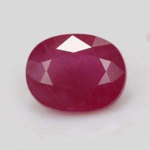 May include: A loose oval-cut ruby gemstone with a deep, saturated magenta-red colour. The stone features a faceted surface with a symmetrical arrangement of triangular and kite-shaped facets that reflect light, creating a clear, polished appearance.