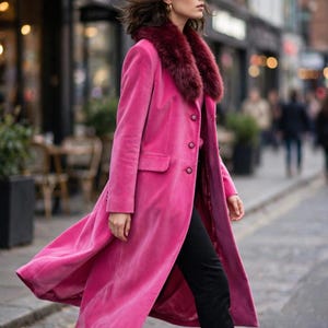 May include: A long, vibrant pink velvet coat featuring a deep burgundy faux fur collar, long sleeves, and a front button closure with three visible buttons. The coat is paired with slim-fitting black trousers and black ankle boots.