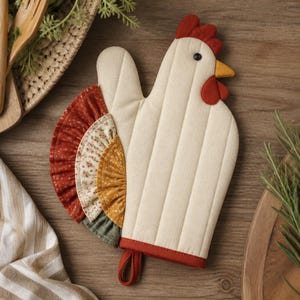 Pode incluir: Uma luva de forno acolchoada em forma de galinha, com corpo cor de creme, crista e barbelas vermelhas, bico amarelo e olho preto. A cauda consiste em camadas de tecido com folhos em tons de ferrugem, creme floral, mostarda e verde azeitona.