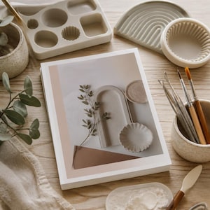 May include: A white book featuring a cover image of beige and brown geometric shapes, a small plant sprig, and circular dishes. Surrounding the book are silicone molds, a ceramic cup with paintbrushes, a small bowl, and a palette knife.