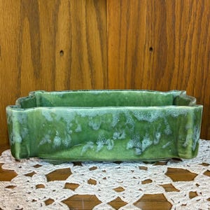 May include: A rectangular green ceramic planter with a scalloped edge and a glossy glaze finish. The surface features a mottled texture with lighter green and white highlights along the top rim and corners, supported by small feet at the base.