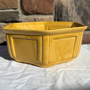 May include: A square yellow ceramic bowl with a glossy finish, featuring raised panel detailing on each side and a wide, flat rim. The bowl has a distinct geometric shape with clean, straight lines and a smooth, reflective surface.