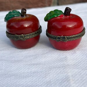May include: Two red apple-shaped trinket boxes with glossy finishes, green leaves, and dark brown stems. Each box features a hinged metal band with an antique brass finish and a small bow-shaped clasp detail at the center of the closure.