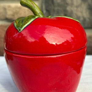 May include: A glossy red ceramic apple-shaped container with a removable lid. The top of the lid features a sculpted green stem and a single green leaf with dark shading, providing a realistic appearance to the fruit-shaped storage vessel.