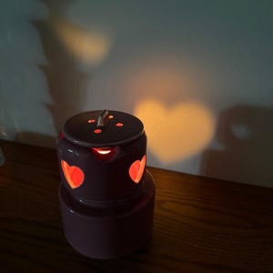 May include: A cylindrical ceramic wax warmer in a muted purple hue, featuring heart-shaped cutouts around the upper section. A removable lid sits on top, and the interior light casts a distinct heart-shaped shadow onto the wall behind it.