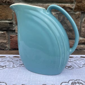 May include: A light blue ceramic pitcher with a glossy finish, featuring a curved silhouette, a wide pouring spout, and a smooth handle. The exterior surface displays subtle, raised concentric arc detailing along the side, while the interior is a contrasting off-white color.
