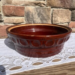 May include: A large, round, deep ceramic mixing bowl with a glossy dark brown glaze. The exterior features a decorative band of raised circular indentations around the upper rim, with horizontal ridges encircling the lower base section.