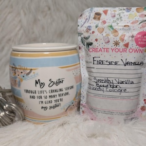 Può includere: Un scaldacandele in ceramica con coperchio, con strisce pastello e dettagli floreali. Il testo sullo scaldacandele recita "My Sister" e un messaggio. Accanto, un sacchetto con l'etichetta "Create Your Own!" con "Fireside Vanilla" e altre note di profumo.