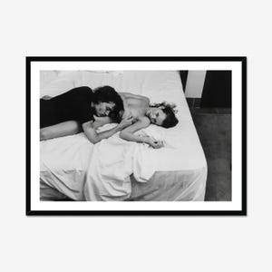 Pode incluir: Uma impressão fotográfica a preto e branco numa moldura preta que retrata duas pessoas deitadas juntas numa cama branca. Uma pessoa veste uma peça escura enquanto a outra é parcialmente visível, ambas sobre lençóis brancos com dobras e texturas.
