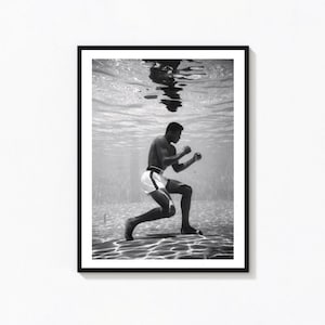 Pode incluir: Uma fotografia emoldurada a preto e branco que retrata uma pessoa debaixo de água numa piscina. A pessoa está numa posição de boxe, vestindo calções brancos, com o seu reflexo visível na superfície da água. A cena captura padrões de luz.