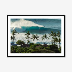 Op de afbeelding: Een ingelijste landschapsfoto met een grote, turquoise oceaangolf die breekt nabij een tropische kustlijn. Hoge palmbomen en donkere daken zijn zichtbaar op de voorgrond, terwijl verschillende surfers in het water liggen.
