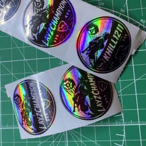 May include: A sheet of five circular holographic stickers featuring a black graphic of a motorcycle and rider. The text LAYZCHAMPION and KHILL1211 is printed on the stickers. The stickers have a rainbow metallic finish and are arranged on a white backing.