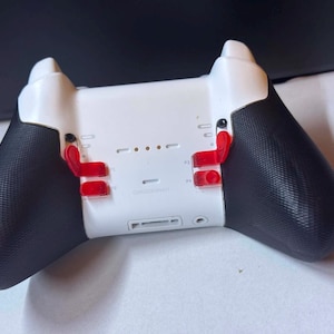 May include: A white video game controller featuring black textured grips on the handles. The back of the device includes four translucent red paddle buttons, a central charging port, and a small headphone jack located at the bottom center.