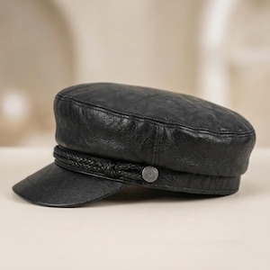 May include: A black cap with a textured surface, featuring a short visor and a braided cord detail across the front. The cord is secured on each side by a small, circular metallic button embossed with the letter U, creating a structured look.