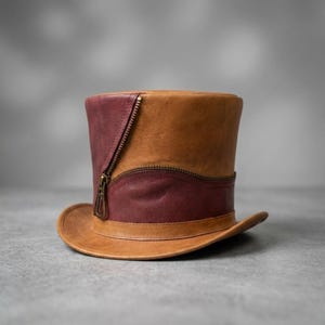 May include: A top hat constructed from tan and maroon leather panels. The hat features a curved metal zipper detail that runs vertically and horizontally across the side, with a matching tan leather brim and a structured, flat-topped cylindrical crown.