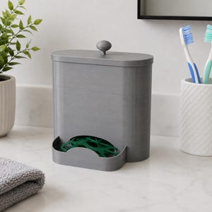 May include: A grey, oval-shaped plastic dispenser with a removable lid featuring a small knob handle. The front of the container has a curved cutout at the base, revealing a stack of green dental floss picks stored inside the unit.
