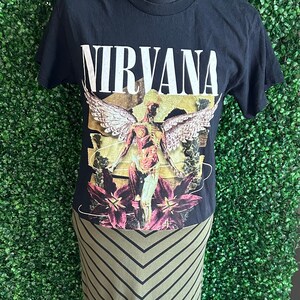 May include: Black t-shirt with the word "NIRVANA" in white block letters. The shirt features a graphic of a winged figure with floral accents. Paired with an olive green and black chevron striped skirt.