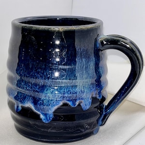 May include: A ceramic mug with a ribbed, cylindrical body and a curved handle. The exterior features a deep navy blue base glaze with a vibrant, drippy cobalt blue and white glaze effect cascading from the rim down the sides of the vessel.
