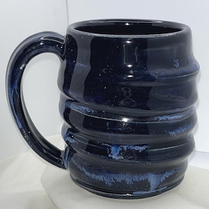 May include: A dark navy blue ceramic mug with a glossy finish and a series of horizontal ridges along the body. The mug features a large, curved handle on the left side and subtle lighter blue streaks that highlight the textured, ribbed surface.